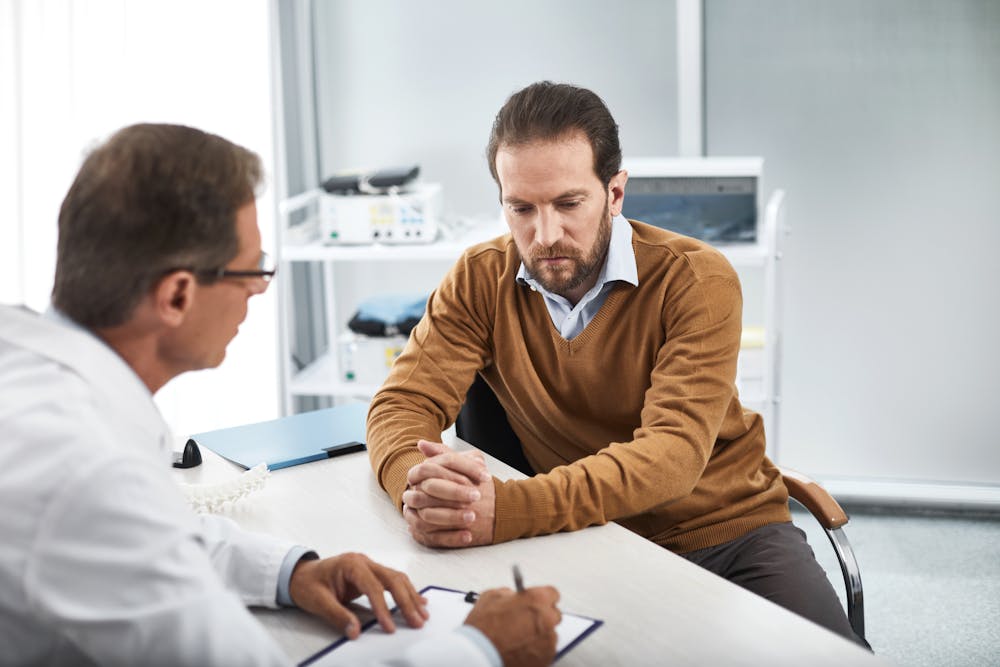 A man consults with a doctor across a desk in a well-lit, clinical room; medical equipment is visible in the background. Both individuals appear engaged in conversation. - The Law Offices Of Justin Frankel